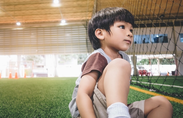 サッカーのスポーツフィールドに座っている小さな男の子