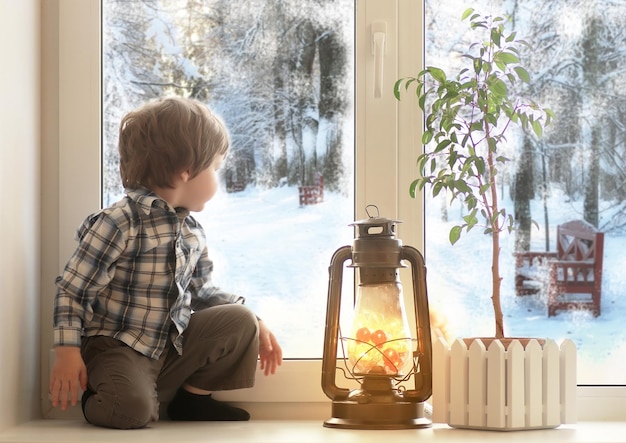 Little boy sitting on a white window sill and looks out the window at winter landscape