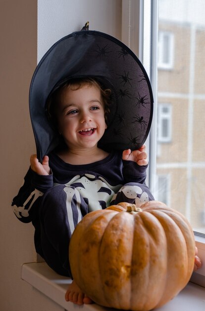 A little boy in skeleton costume and witch hat for halloween is sitting at the window and laughiting