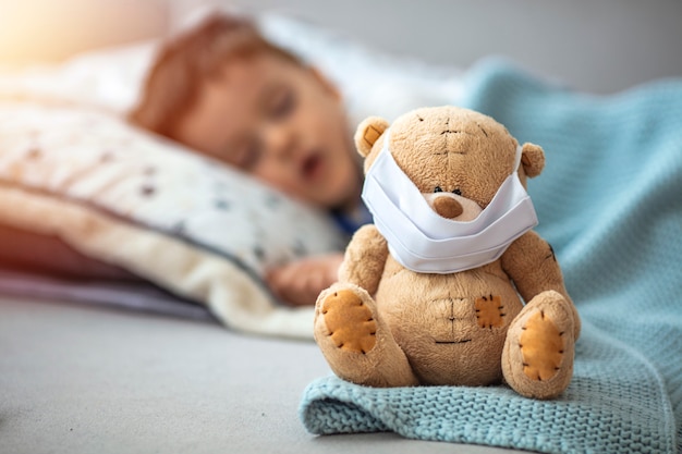 Photo little boy sleeping in his bed with his teddy bear wearing a mask to protect him against corona virus covid-19 / 2019-ncov concept