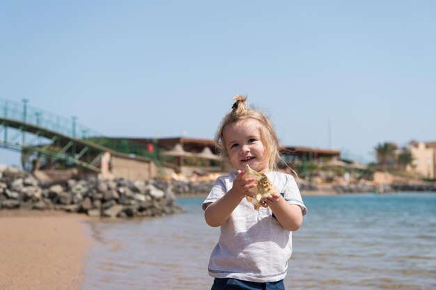 Улыбка маленького мальчика с ракушкой на морском пляже Детская игра с ракушкой на солнечном морском пейзаже Открытие свободы и приключения Летние каникулы на море Концепция счастливого детства