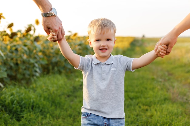 Little boy smiling and holding his parents hands on the background of countryside Kid with family