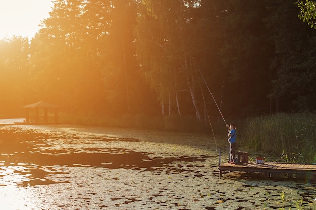 Little boy standing and fishing on a wooden dock at the sunset.