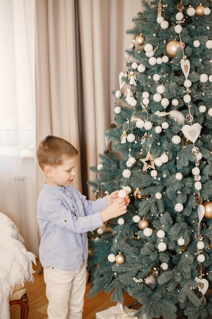 Little boy standing on the floor near christmas tree at home. Boy is hanging Christmas tree decoration. Boy wearing blue shirt and beige trousers.