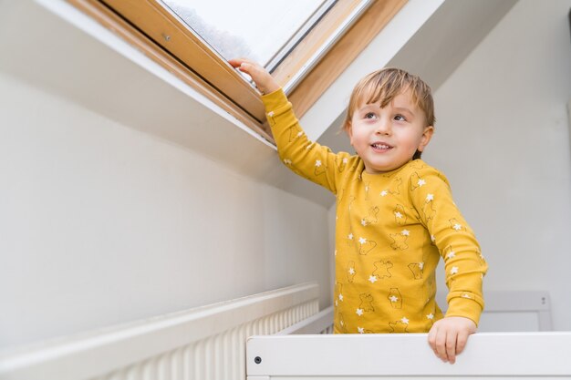 A little boy standing in his bed in the morning and touching roof window by his hand.