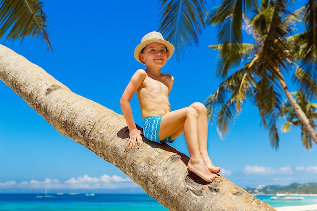 Маленький мальчик в соломенной шляпе с удовольствием на кокосовой пальме на песчаном тропическом пляже. Концепция путешествий и семейного отдыха.