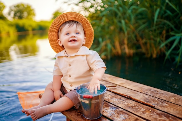 木製のドックの端に座って、日没時に湖で釣りをしている麦わら帽子の少年。