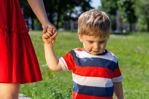 A little boy in a striped T-shirt walks with his mother's hand in the park