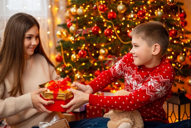 飾られたクリスマスツリーの下に座ってプレゼントのために戦う小さな男の子と10代の少女の兄弟クリスマスのお祝い