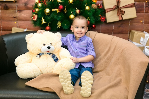 A little boy of three years old is sitting on a leather sofa holding a soft bear in his hands in a New Year's atmosphere.