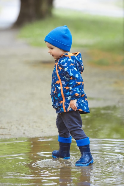 Little boy in waterproof blue rubber boots in puddles with splash after the rain