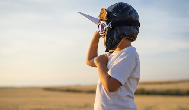 ヘルメットをかぶった少年と日没時に紙飛行機をプレイしながら飛行士になることを夢見る