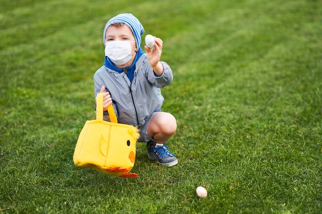 Little boy wearing protective mask hunting for Easter egg in spring garden during coronavirus pandemic