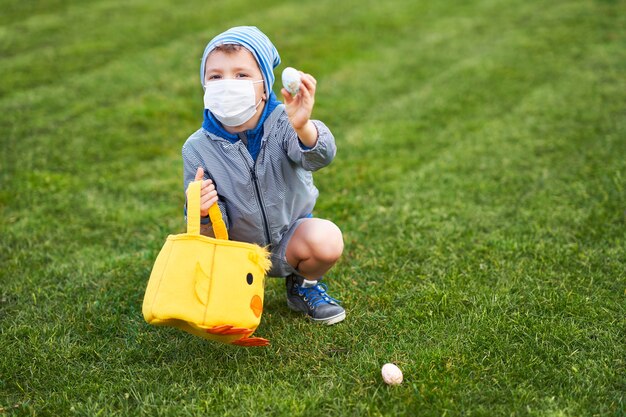 Little boy wearing protective mask hunting for easter egg in spring garden during coronavirus pandemic