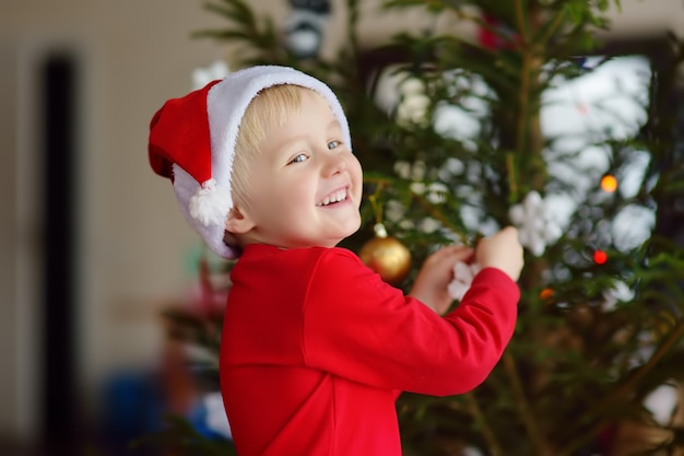 クリスマスを祝うために準備ができているサンタ帽子をかぶっている少年。ガラスのおもちゃでクリスマスツリーを飾るかわいい子