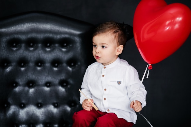 Little boy in white shirt and red trousers sitting on the armchair with red heart balloon on the  Valentines day