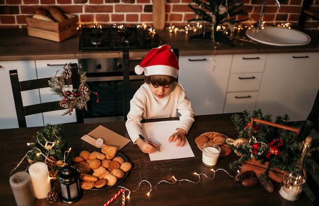 A little boy in a white sweater and a New Year's hat is sitting at a table and making a list of gifts for Christmas