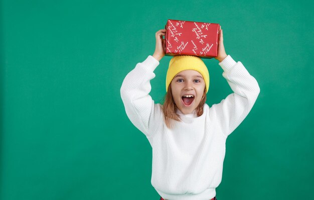 A little boy in a white sweater and a yellow hat is holding a Christmas gift over his head , shouting loudly and emotionally with his mouth open .