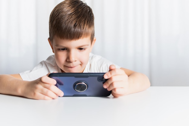 흰색 티셔츠를 입은 어린 소년이 집에서 전화로 게임을 합니다. 행복한 아이가 스마트폰을 보고 있습니다.