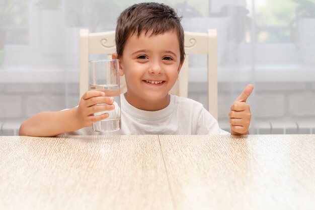 a little boy in a white Tshirt at home holds a glass of water in his hands