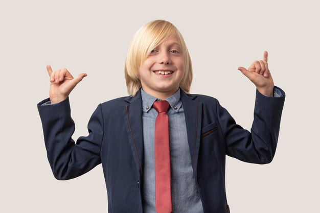 Little boy with blond hair in blue jacket and red tie smiling and pointing fingers up make shaka gesture while standing against white background