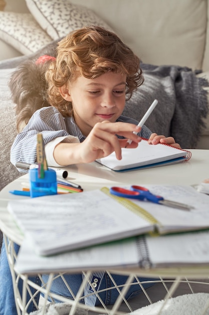 Little boy with curly blond hair in casual clothes sitting at table and taking notes in notepad in living room at home during weekend