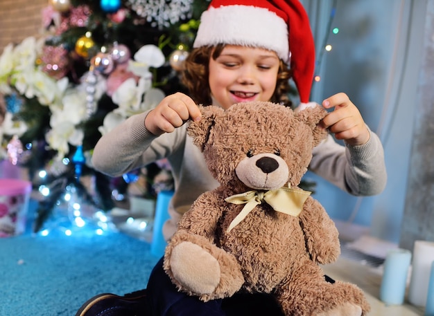 A little boy with curly hair in a red Santa hat holding a Teddy bear