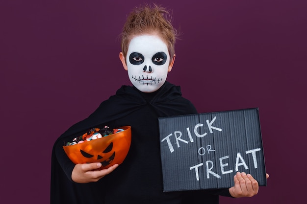 사진 할로윈 메이크업과 해골 의상을 입은 어린 소년이 과자와 칠판을 들고 있다: trick or treat. 할로윈 개념입니다.