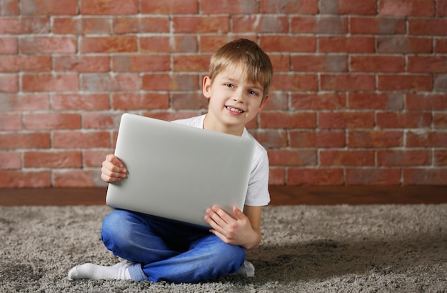 Photo little boy with laptop on fur carpet against brick wall background