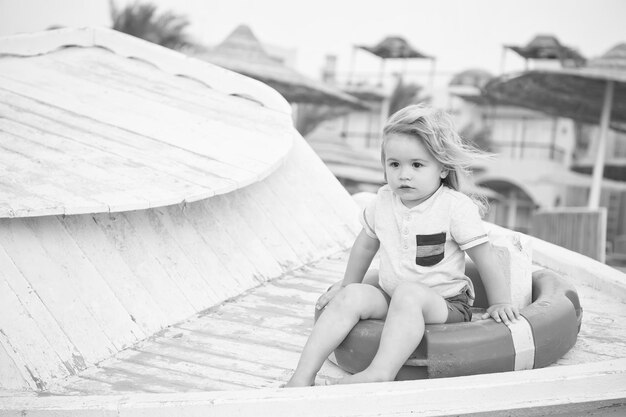 Little boy with lifebuoy on tropical beach Child sit in ring buoy on sunny day Kid with blond hair have fun outdoor Summer vacation and leisure activity Safety rescue and help concept