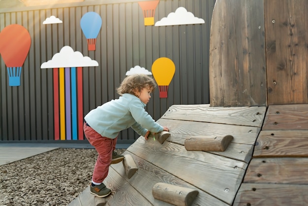 Маленький мальчик с длинными волосами, поднимающийся по деревянной горке на общественной игровой площадке на открытом воздухе