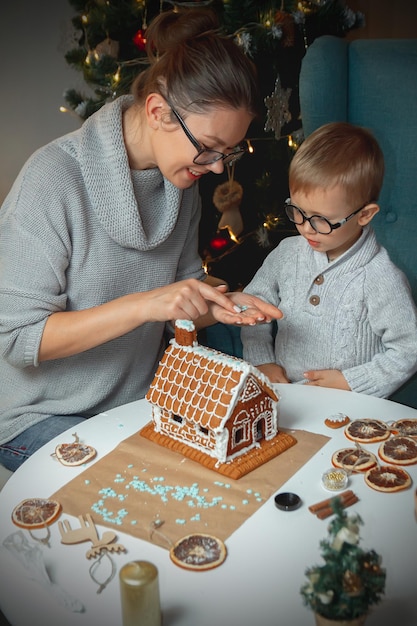 ママと一緒に小さな男の子がクリスマスのジンジャーブレッドの家を一緒に飾る