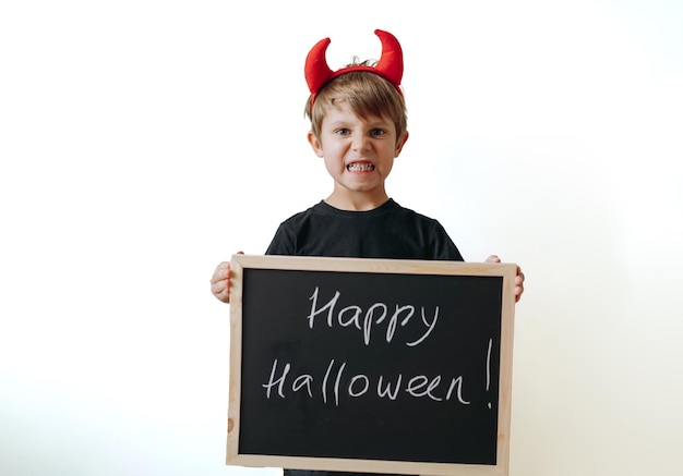 Little boy with red devil horns and black board with Happy Halloween text on white background