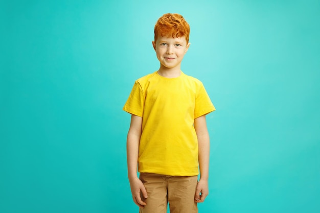 Photo little boy with red hair wearing yellow t shirt standing confidently on blue turquoise isolated