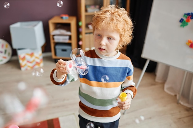 Photo little boy with short ginger hair in striped pullover looking at multiple soap bubble while playing