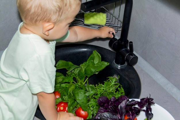 A little boy with a short haircut helps to cook in the kitchen Washes fresh vegetables