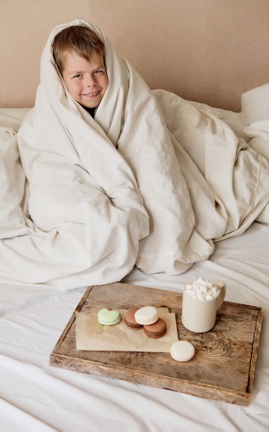 Little boy wrapped in a blanket with breakfast in bed. cocoa with marshmallows and macaroons