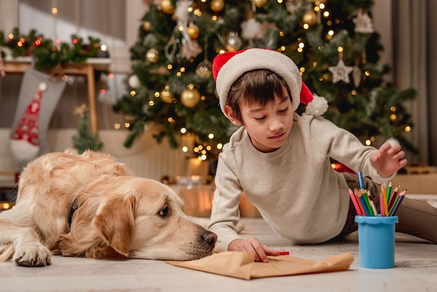 Little boy writing letter to santa while lying beside golden retriever dog on floor at home