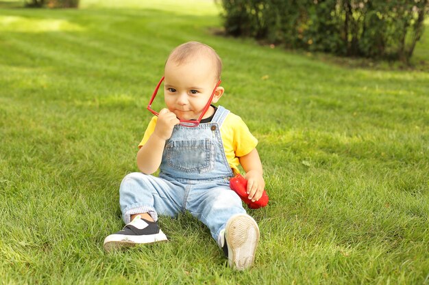 a little boy in a yellow Tshirt is sitting on the lawn in the park holding a red bell