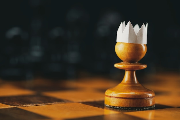 Little brave pawn wearing artificial paper crown suit on chessboard with figures, business entrepreneur leadership concept.