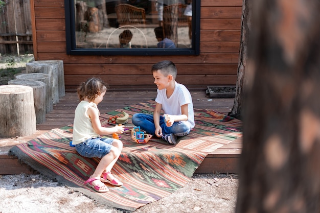 Photo little brother and sister sitting on the porch near a wooden house on an embroidered carpet, playing educational games, playing with a constructor, enjoying the summer