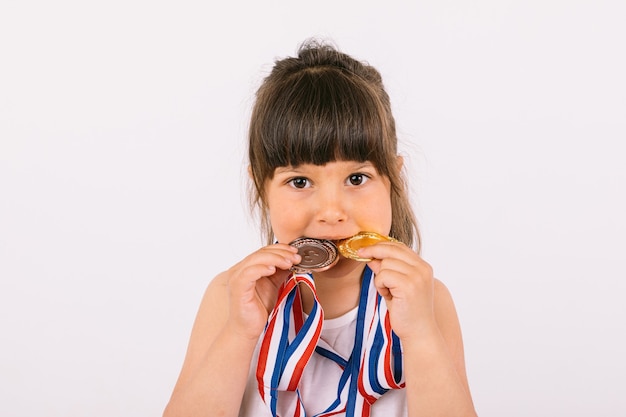 Маленькая шатенка с медалями чемпиона по спорту, кусая их. Концепция спорта и победы