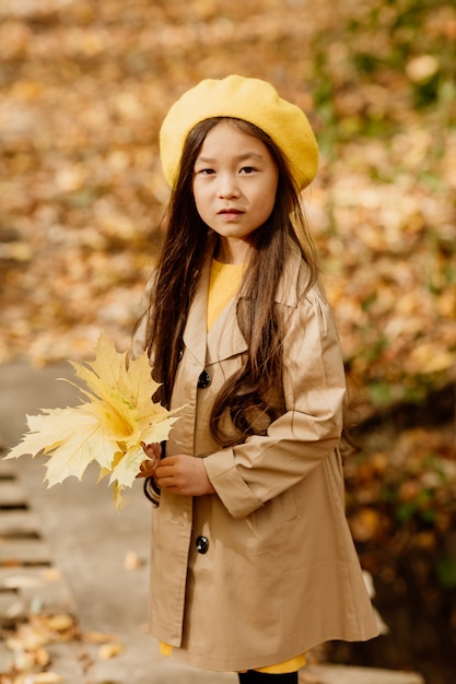 가을 옷을 입은 작은 갈색 머리 아시아 소녀는 개울 근처의 가을 숲에서 산책