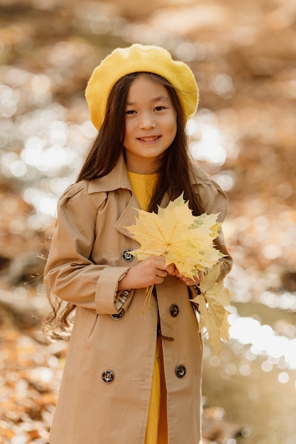 사진 가을 옷을 입은 작은 갈색 머리 아시아 소녀가 개울 근처의 가을 숲을 걷는다
