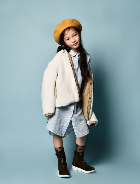 Photo little brunette asian schoolgirl in double sided jacket, shirt dress, brown beret, boots. she smiling, posing against blue studio background. childhood, fashion, hipster style. full length, copy space