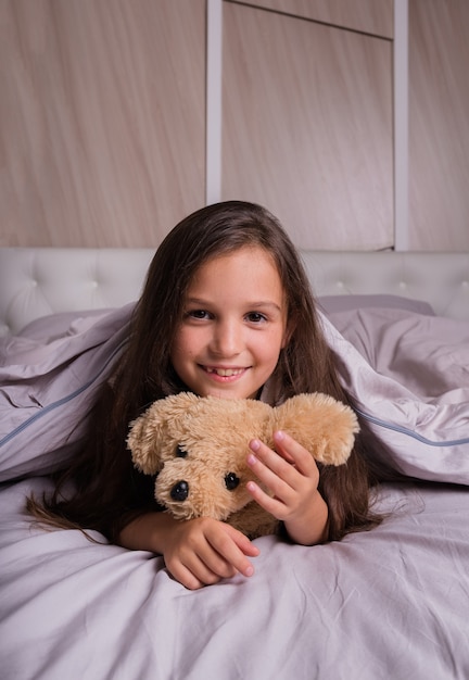 A little brunette girl in pajamas is lying on the bed linen on the bed with a teddy bear