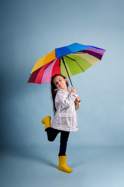 A little brunette girl in a raincoat and rubber boots stands\
with a multi-colored umbrella on a blue background with a place for\
text