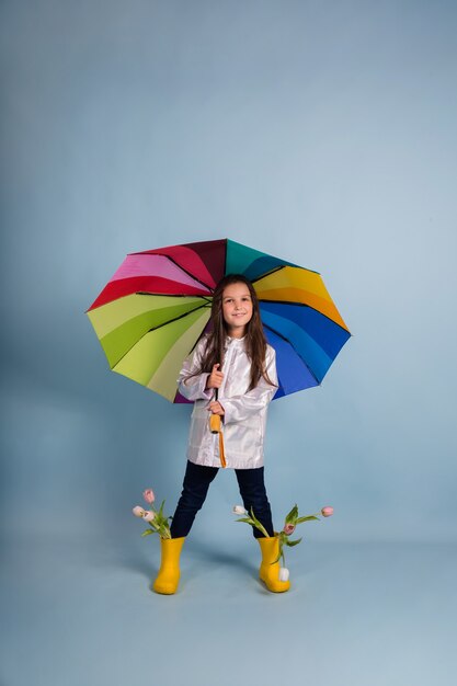 A little brunette girl in a raincoat and rubber boots with flowers stands under a multi-colored umbrella on a blue background with a place for text