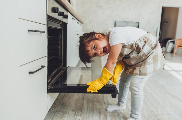 A little brunette girl wipes the oven in the kitchen at home\
cleaning the kitchen a child can have a stove in a white\
kitchen