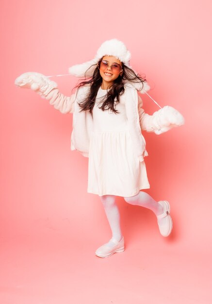 Piccola ragazza bruna con i capelli lunghi in un cappello di pelliccia invernale e guanti su uno sfondo rosa.